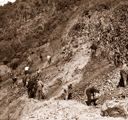 Construction of Great Ocean Road. Workers carve the start of track across a steep slope with pick & shovel. A team before has cleared the vegetation. Distant and near views available. c1920s
