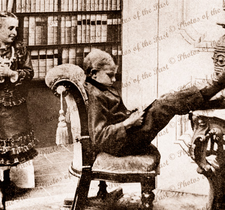 A man of learning. Boy with feet up reading a book. Admirer looks on. c1900