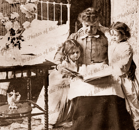 Bedtime reading. Grandmother(?) reads to two small girls in night attire. c1900